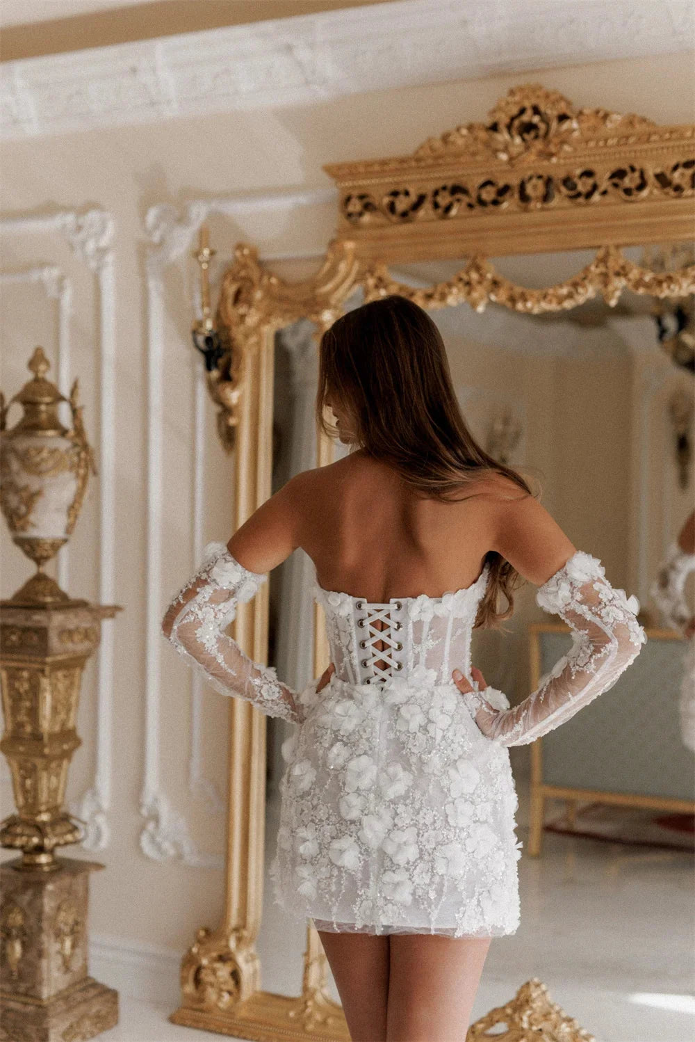 Woman in a white lace dress standing in front of an ornate mirror.