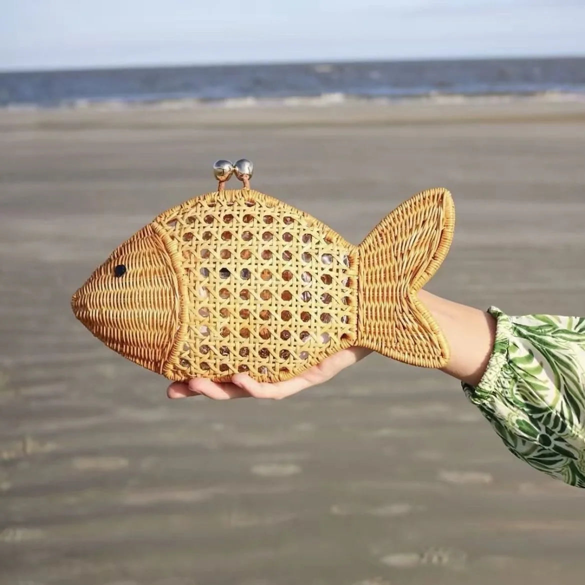 Woven fish-shaped handbag held by a person on a beach.