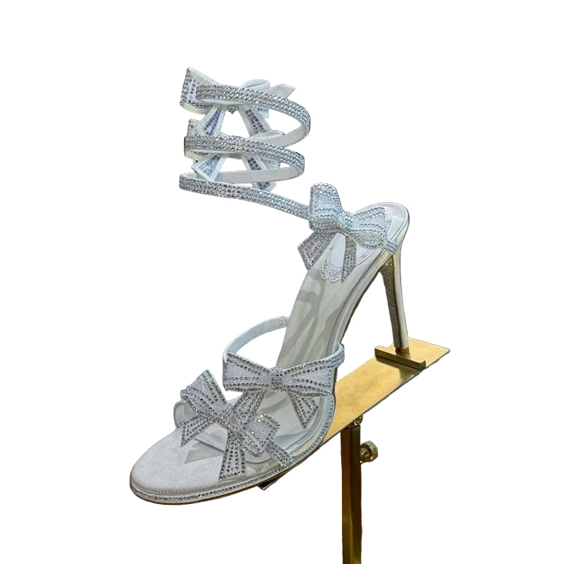 Silver high-heeled sandals with bow details on a stand against a textured wall.