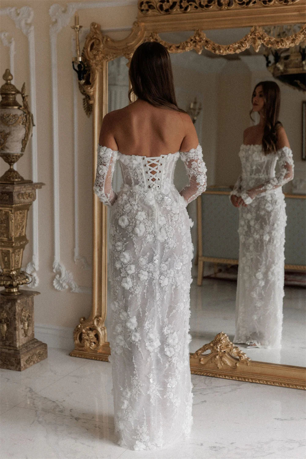 Woman in a white lace dress standing in front of a large ornate mirror.