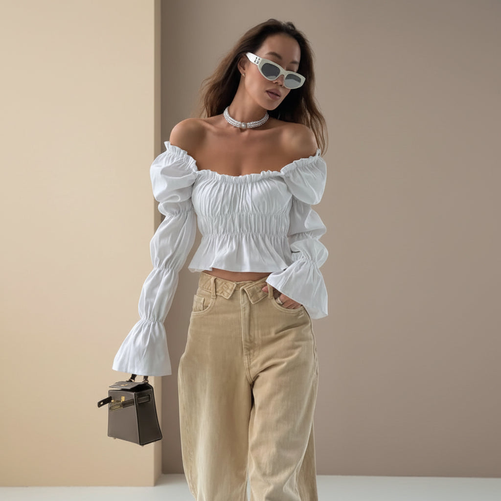Woman wearing a white off-shoulder top and beige pants with sunglasses, standing in a modern architectural setting.